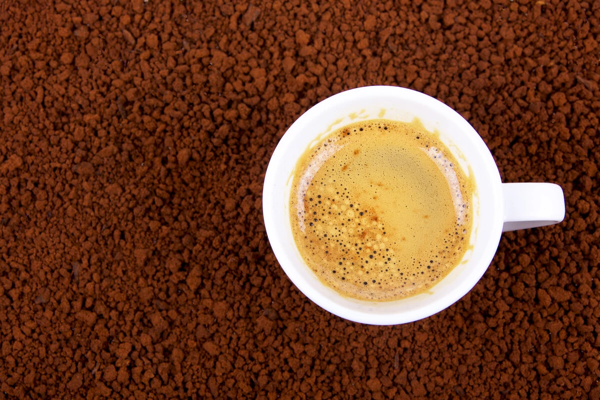 Tazzina bianca di espresso con crema dorata e bollicine, vista dall'alto su un letto di granuli di caffè marrone